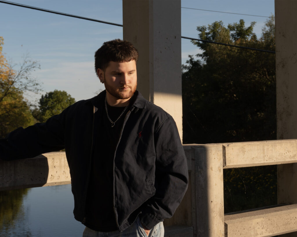 A young man with short dark hair and a trimmed beard stands on a concrete bridge during golden hour. He wears a dark jacket over a black shirt and a chain necklace, resting one arm on the railing while looking off to the side. Warm sunlight casts soft shadows across his face, with trees and calm water visible in the background.