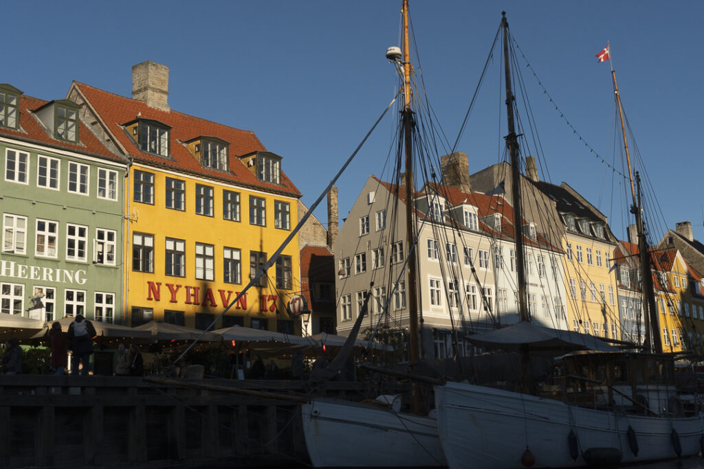 Colourful multi-story buildings with red-tiled roofs line a waterfront canal. The facades are painted green, yellow, and beige, with large windows. In the foreground, wooden boats are docked along the quay, and a Danish flag flies from one mast against a clear blue sky.
