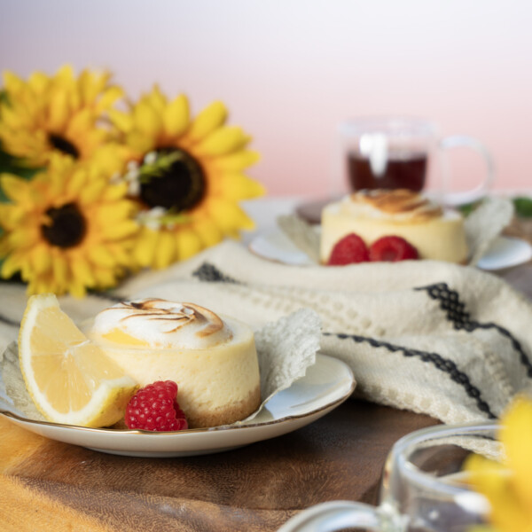 Two small lemon merange cheese cakes plated with a slice of lemon and raspberries and sat on top of a wooden pallet. A clear teacup and a bundle of sunflowers sit in the background.