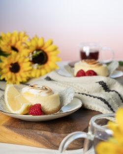 Photo Two small lemon merange cheese cakes plated with a slice of lemon and raspberries and sat on top of a wooden pallet. A clear teacup and a bundle of sunflowers sit in the background.